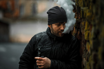 Portrait of a bearded young white guy in the modern hat and black jacket standing near old destroyed red brick wall. Lifestyle.