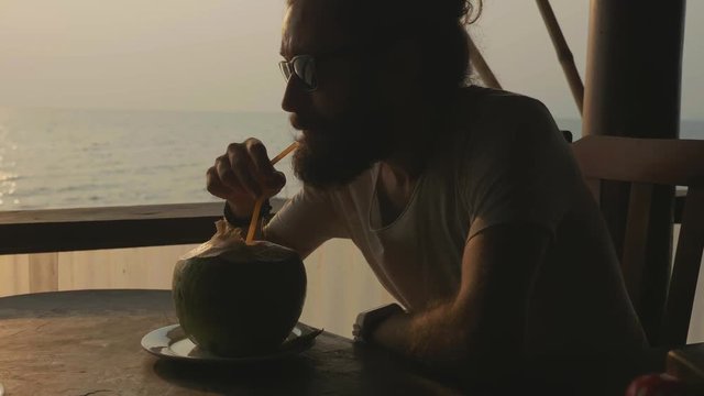 Bearded man sits at seaview cafe and drinks coconat water in slow motion