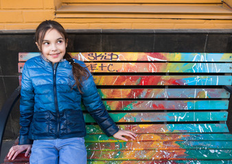 Beautiful girl sits on a painted colored bench and smiles