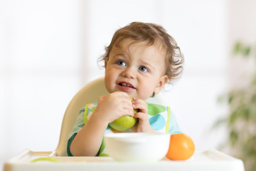 Smiling little kid child baby boy toddler sitting in highchair and eating big green apple fruit portrait indoors
