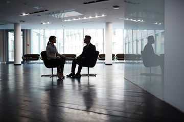 Fototapeta na wymiar Confident young manager sitting opposite her male partner while brainstorming on joint project, interior of spacious dim office lobby on background