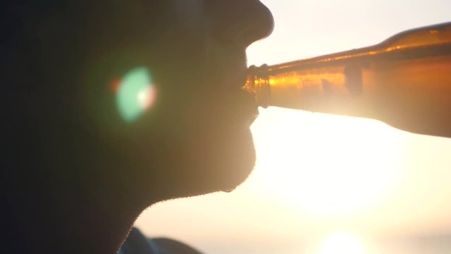 A man drinks beer from a cold bottle, against the background of a sunset with the effect of a lens, drops roll over his beard. slow motion. 1920x1080. full hd