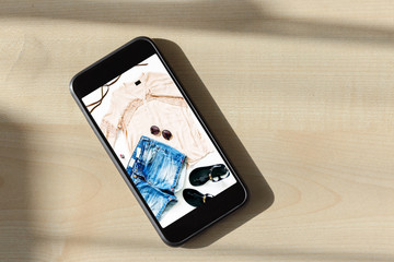 Online shopping concept. Smartphone with photo Summer set of female clothes. Female denim shorts with blouse and sandals on the mobile phone screen, top view, flat lay