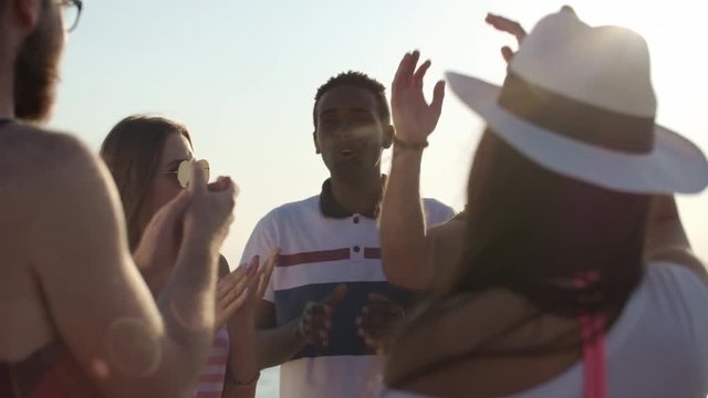 Medium shot of happy multiethnic group of friends clapping and celebrating something on hot summer day on beach