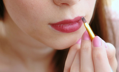 Naklejka premium Closeup of a woman's hand with a brush that puts bright lipstick on her lips.