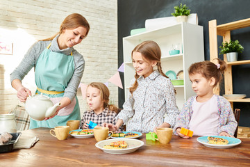 Smiling young woman wearing apron pouring tea in cups while her adorable little daughters looking at homemade pastry with admiration, interior of dining room on background