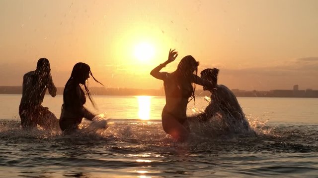 PAN of silhouettes of happy young friends playing in lake and splashing water at each other at sunset
