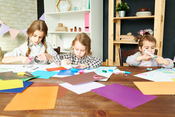 Fototapeta premium Three little sisters wrapped up in creating pictures from colored paper while gathered together at modern living room