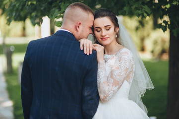 A beautiful bride in a lace dress leaned on the groom's shoulder. Newlyweds in a park with green grass.