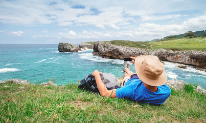 Fototapeta premium Backpacker traveler rests on the rocky sea side and take mobil photo of beautiful sea landscape