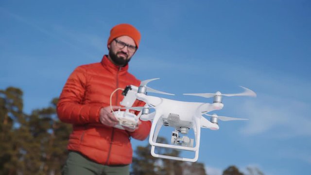Male hipster in red hat and jacket launches a quadrocopter in an open space. Man in glasses controls the copter, using radio-controlled control panel. Operator makes an areial photo using a drone