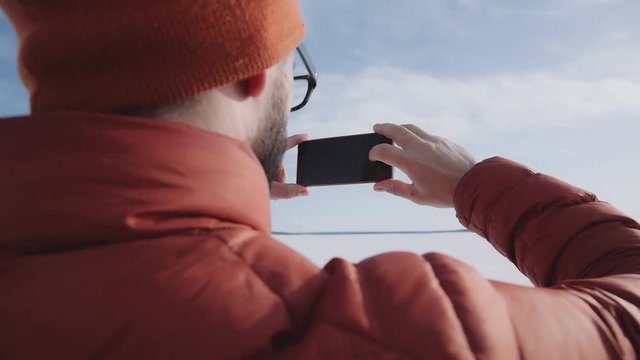 A traveler in a bright jacket stands against the sun and takes pictures of the winter nature on the smartphone. Hicker takes a photo on the phone: the admiration of nature on a sunny day.