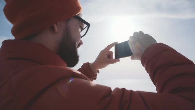 A traveler in a bright jacket stands against the sun and takes pictures of the winter nature on the smartphone. Hicker takes a photo on the phone: the admiration of nature on a sunny day.