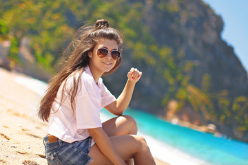 Young happy teenage girl hipster sits on the sand in shorts and rests surrounded by beach and green mountains and smiles at the hotel Oludeniz, Fethiye district in Turkey,