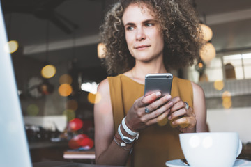 Pretty curly woman in city cafe, with cup of coffee and laptop, typing using mobile phone