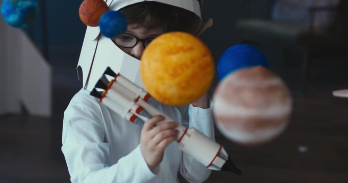 CU Cute little boy wearing cardboard astronaut helmet flying toy rocket through planets, exploring deep space. 4K UHD 60 FPS SLOW MO