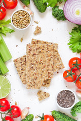 Top view of whole grain rye crispbreads surrounded fresh vegetables, greens and seeds over white background. The concept of organic and vegetarian food.