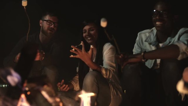 Tilt down of cheerful young friends sitting by campfire at night and chatting while roasting marshmallows