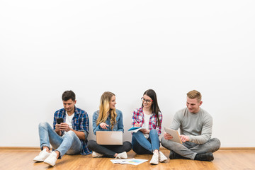 The happy friends with gadgets sit on the floor on the white wall background