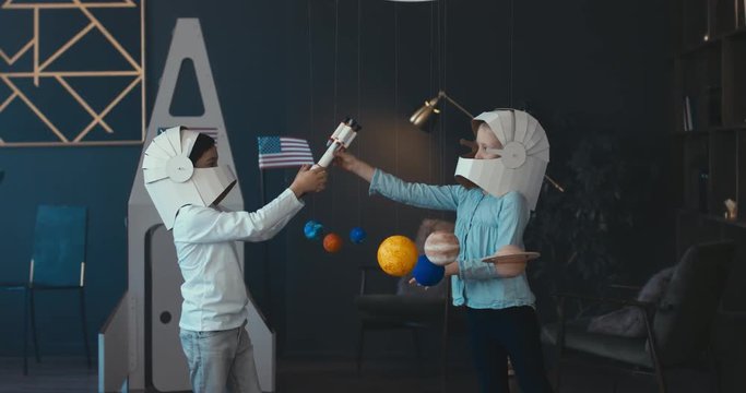 WIDE Cute little boy and girl siblings or friends wearing cardboard astronaut helmets flying toy rocket through planets, exploring deep space. 4K UHD 60 FPS SLOW MO