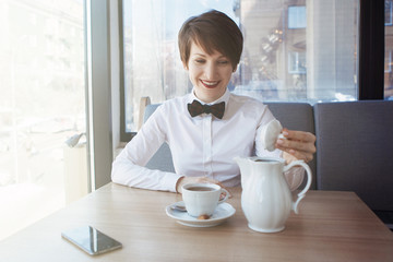 A young pretty girl in a white blouse with a bow tie at a table in a cafe smiles, dreams, pours tea from a teapot into a cup