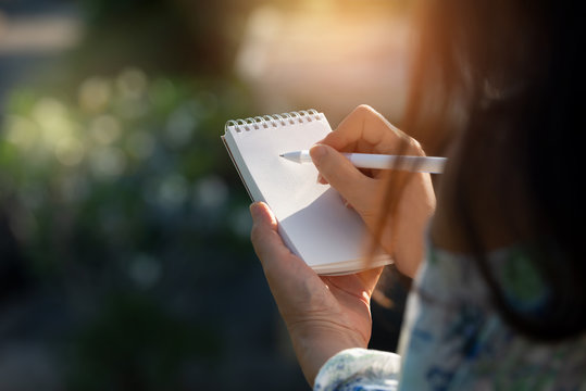 Hand writing on note pad.Woman with long hair hands holding  blanknote writing with white pen  relaxing  by the pool at sunset ,water bokeh  background.