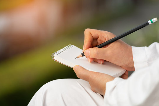 Hand writing on note pad.Woman hands holding  blank note  writing with black pencil relaxing in garden backyard at sunrise ,natural blurred background.