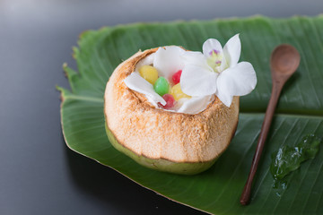 Fototapeta na wymiar Thai traditional dessert.Tab tim grob on ice cube serve in fresh green coconut shell decorated by white plumeria coconut meat wooden spoon and banana leaf on black background.