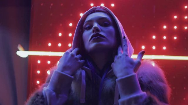 Close-up of beautiful young female face looks at camera in neon light red, winks and shows come with me simbol by finger