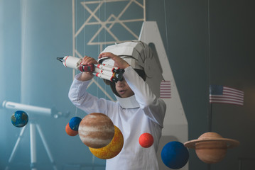 Cute little boy wearing cardboard astronaut helmet flying toy rocket through planets, cardboard spaceship rocket in the background © supamotion