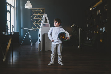 Cute little dreamer boy posing with a helmet at home, pretending to be an astrounaut, cardboard space rocket in the background © supamotion
