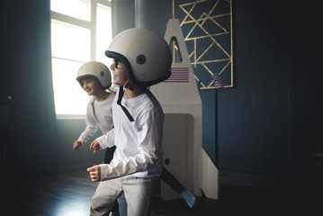 Cute little dreamer siblings boy and girl wearing space helmets pretending to be astronauts on Moon, getting out cardboard space rocket at home