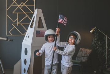 Cute little dreamer siblings wearing space helmets pretending to be astronauts on Moon, placing US flag near cardboard space rocket at home