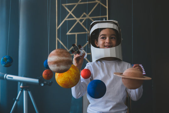 Cute little boy wearing cardboard astronaut helmet flying toy rocket through planets, cardboard spaceship rocket in the background