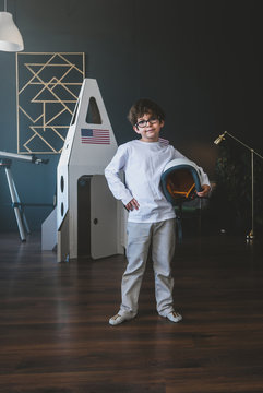Cute little dreamer boy posing with a helmet at home, pretending to be an astrounaut, cardboard space rocket in the background