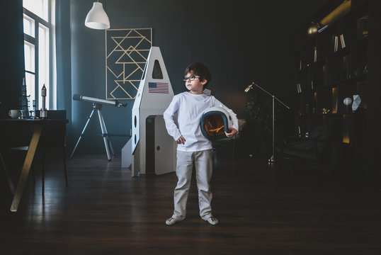 Cute little dreamer boy posing with a helmet at home, pretending to be an astrounaut, cardboard space rocket in the background
