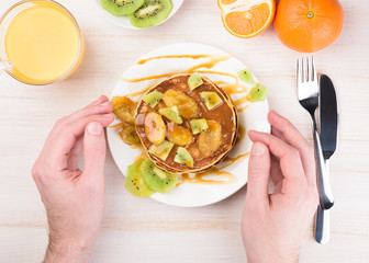 Homemade breakfast on a wooden white table. Stack of pancakes with a grilled bananas and a caramel. Glass of juice, orange, kiwi slices. Female hands, flat lay, top view