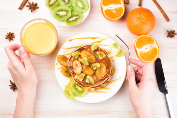 Homemade breakfast on a wooden white table. Stack of pancakes with a grilled bananas and a caramel. Glass of juice, orange, kiwi slices. Female hands, flat lay, top view