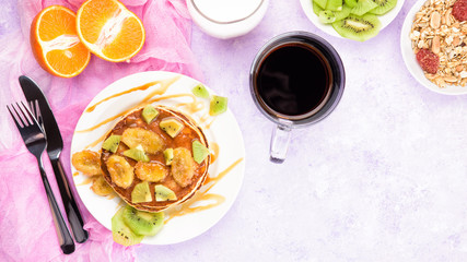 Homemade breakfast on a marble violet table. Stack of pancakes with a grilled bananas and a caramel. Glass of juice, orange, kiwi slices.Healthy breakfast concept, flat lay, top view