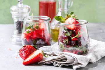 Beetroot and strawberry salad served with arugula and nuts in glass jars with cloth, pepper and bottles of fruit ocet and olive oil over grey table with green wall as background. Healthy eating