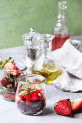 Beetroot and strawberry salad served with arugula and nuts in glass jars with cloth, pepper and bottles of fruit ocet and olive oil over grey table with green wall as background. Healthy eating
