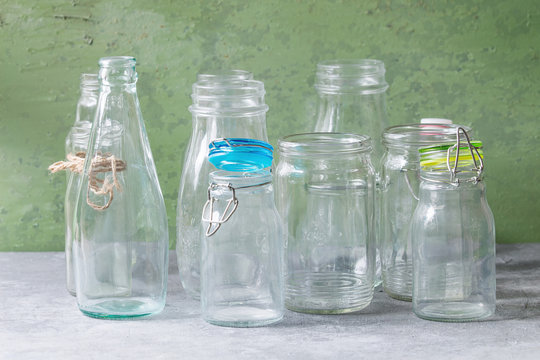 Variety of different shape empty opened glass bottles with and without lids standing on grey table with green wall as background.