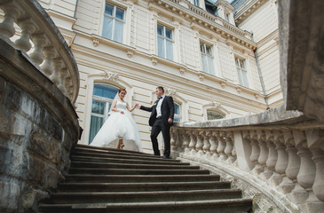 Wedding couple is walking down the stairs in old city. Groom and bride in lace satin dress are near the ancient stone walls palace. Sunny love story in the medieval town.