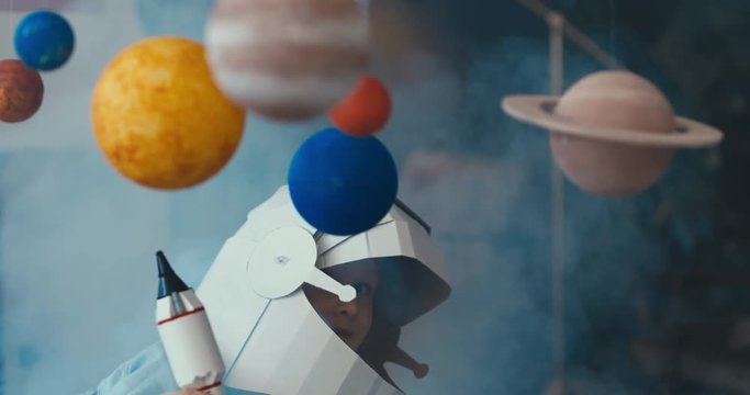 CU Cute little girl wearing cardboard astronaut helmet launching a toy rocket from a spaceport through planets. Imagination concept. 4K UHD 60 FPS SLOW MO