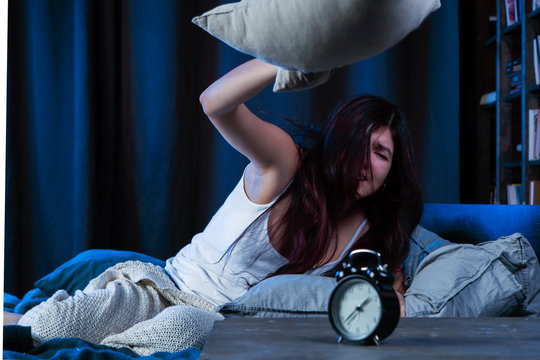 Portrait of dissatisfied woman with insomnia throws pillow sitting on bed next to alarm clock