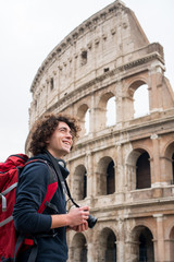 Fototapeta na wymiar Handsome young tourist man with curly hair with a camera and backpack taking pictures of Colosseum in Rome