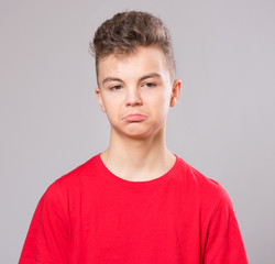 Emotional portrait of caucasian upset problem teen boy. Sad boy looking at camera. Worried  child wearing red t-shirt, on gray background.