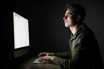 Concentrated young man sitting at the table and using computer in dark room