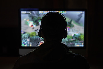 Back view of concentrated young gamer in headphones and glasses using computer for playing game at...