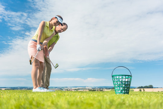Full length side view of a young woman holding an iron club, while exercising the golf swing helped by her experienced instructor outdoors on green grass